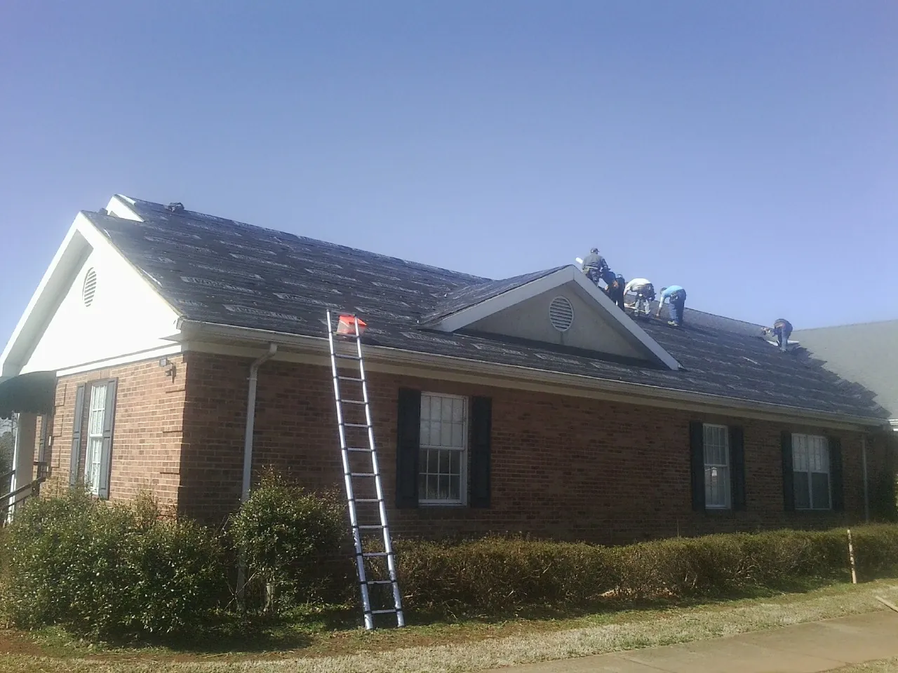 Crew tearing shingles off a brick ranch roof with a ladder set — Emergency Roof Repair work by our crew in Lawrence